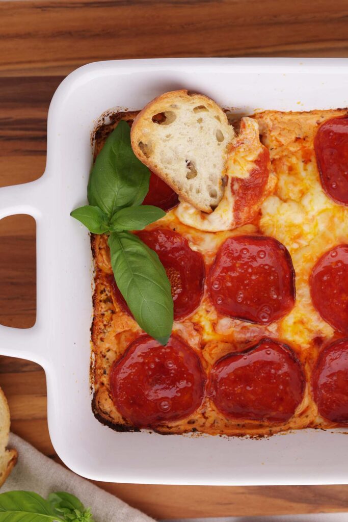 Top down image showing a close up of a white rectangle dish filled with melty cheese that is topped with pepperoni, fresh basil, and crostini