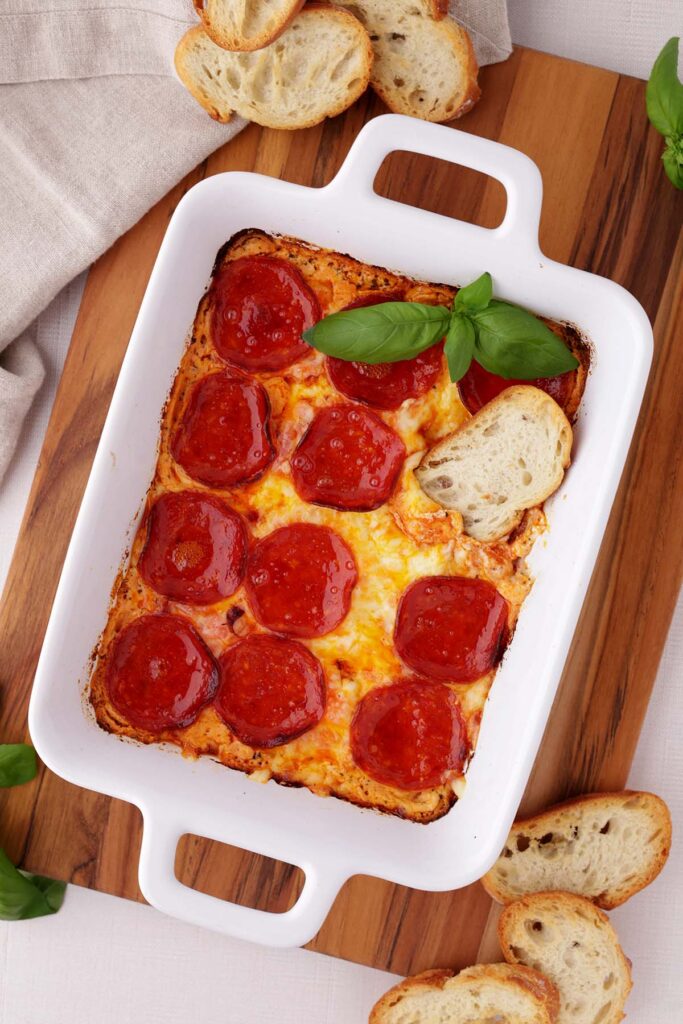 Top down image showing a close up of a white rectangle dish filled with melty cheese that is topped with pepperoni, fresh basil, and crostini