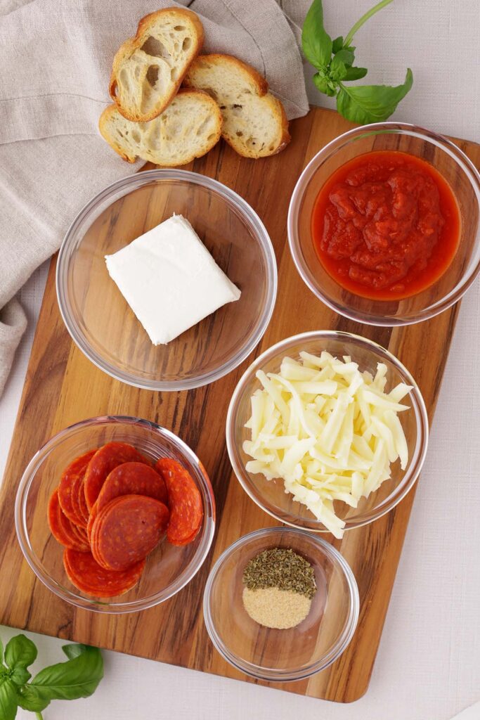 Top down image showing a glass bowl with cream cheese, pizza sauce, pepperoni, mozzarella cheese, and seasonings on a wooden cutting board with crostini and basil on the sides