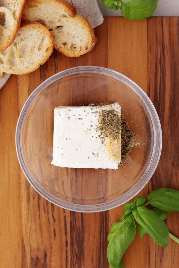 Top down image showing a small glass bowl sitting on a dark wooden table top with cream cheese and seasonings unmixed inside