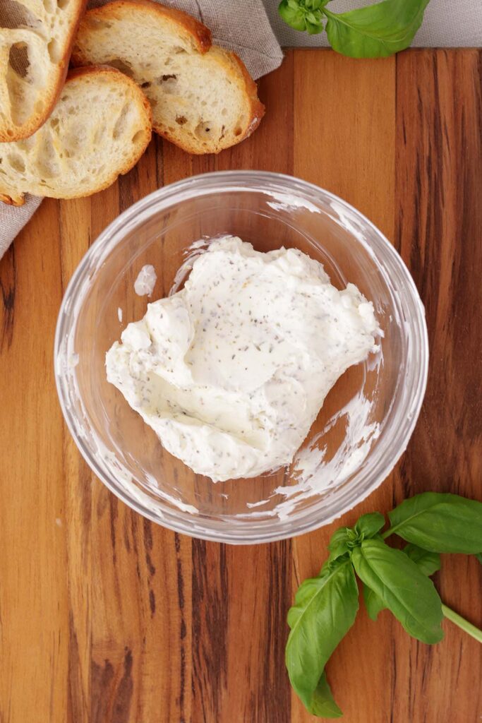 Top down image showing a small glass bowl sitting on a dark wooden table top with cream cheese and seasonings mixed inside