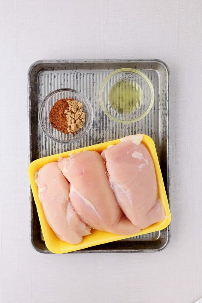 top down image showing chicken breast on a yellow tray with a small glass bowl filled with oil and a second glass bowl filled with seasonings and brown sugar on a metal tray