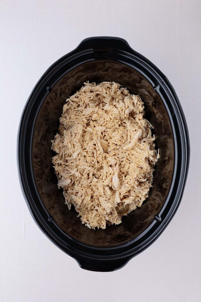 Top down image showing the inside of a black slow cooker filled with shredded chicken