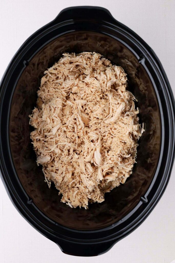 Top down image showing the inside of a black slow cooker filled with shredded chicken
