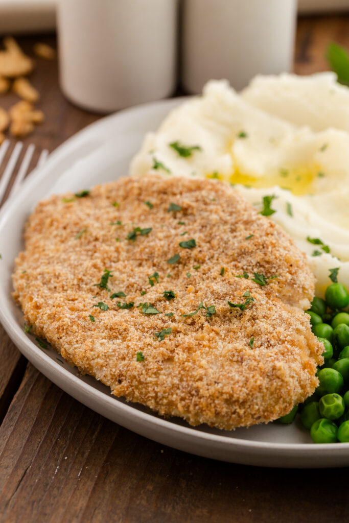 close up image showing a crispy chicken breast on a plate with mashed potatoes and peas