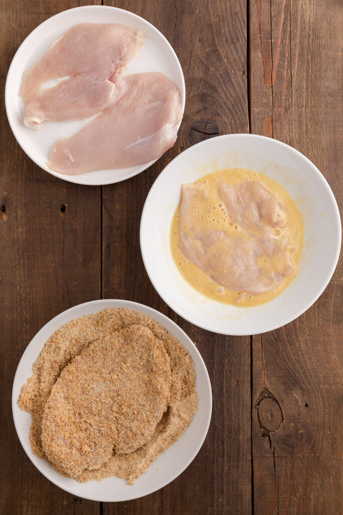 Top down image showing three white plates. The first has pounded chicken breast, the second shows chicken in an egg wash mix, the third has chicken in breadcrumbs