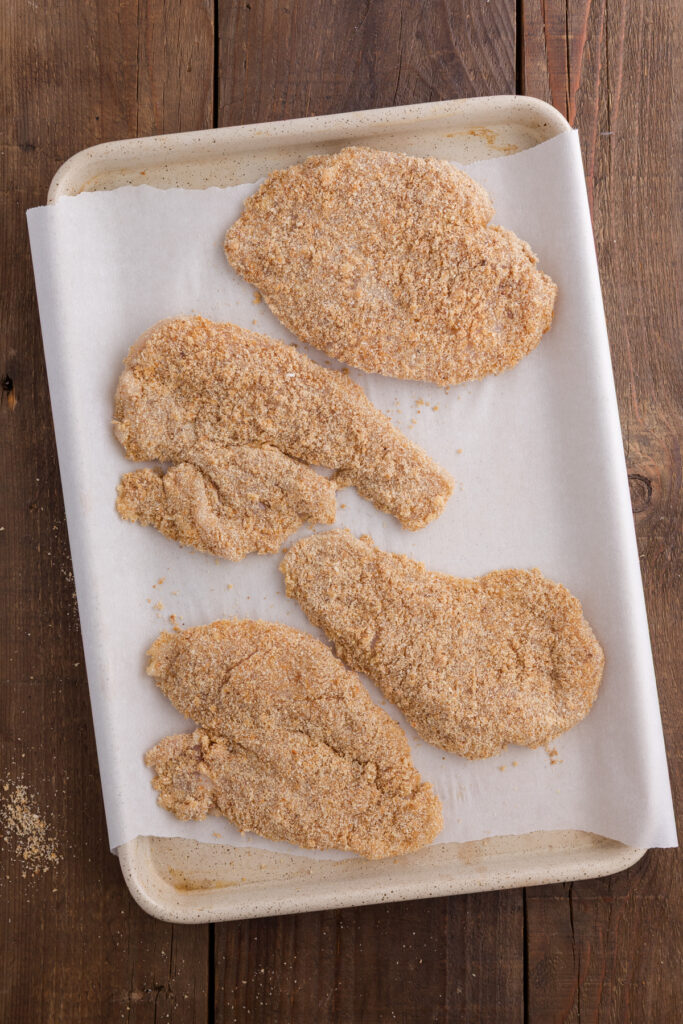 TOp down image showing a parchment lined baking tray layered with uncooked coated chicken breasts