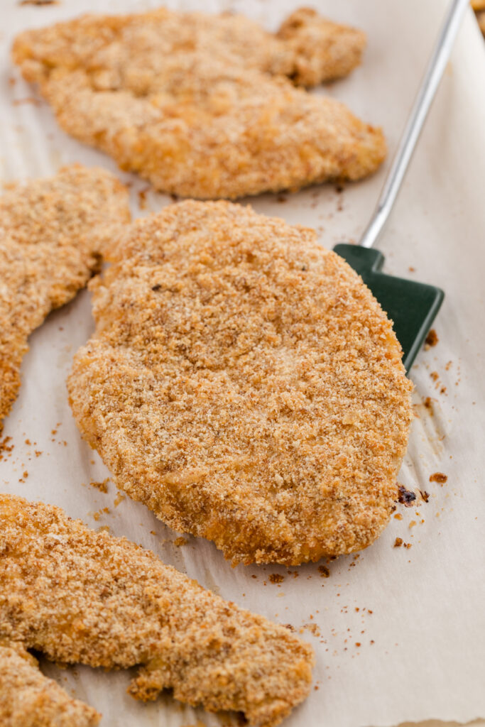 close up image showing crispy baked chicken on a parchment lined tray with one being lifted by a spatula