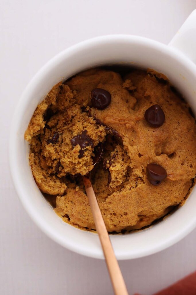 close up image showing a white mug filled with an orange cake topped with chocolate chips. A rust colored napkin is in the background