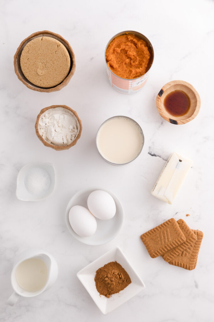 top down image showing pumpkin  puree, brown sugar, flour, condensed milk, salt, eggs, vanilla, butter, cookies, cinnamon and cream on a white table
