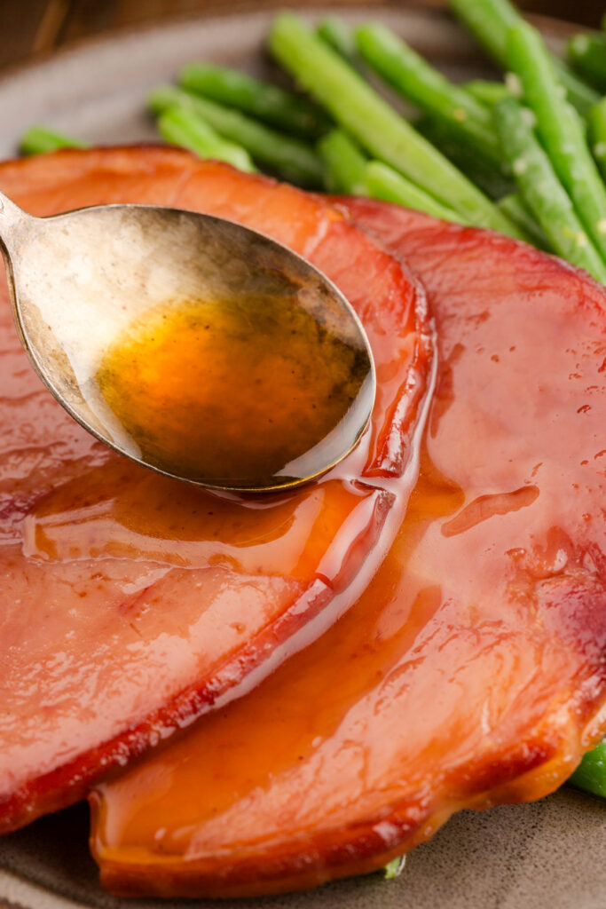 a spoon pouring glaze over the top of ham slices on a plate with green beans
