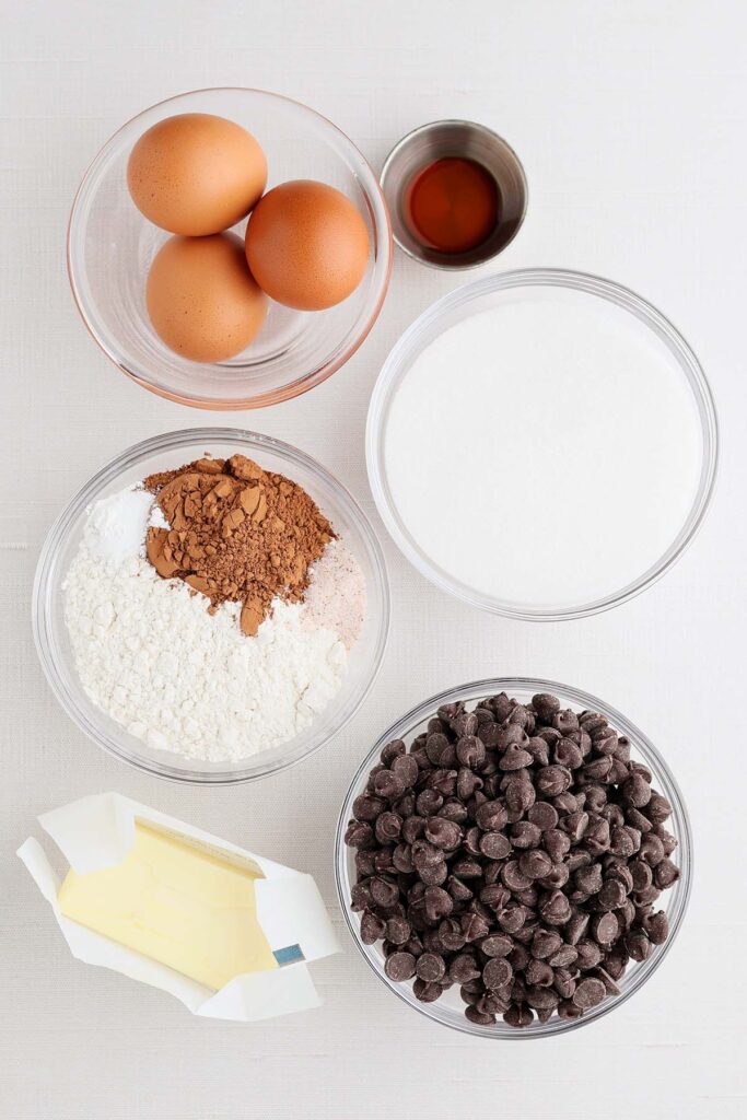 Overhead view of brownie cookie ingredients arranged in bowls, including eggs, sugar, cocoa powder, flour, chocolate chips, butter, and vanilla extract.