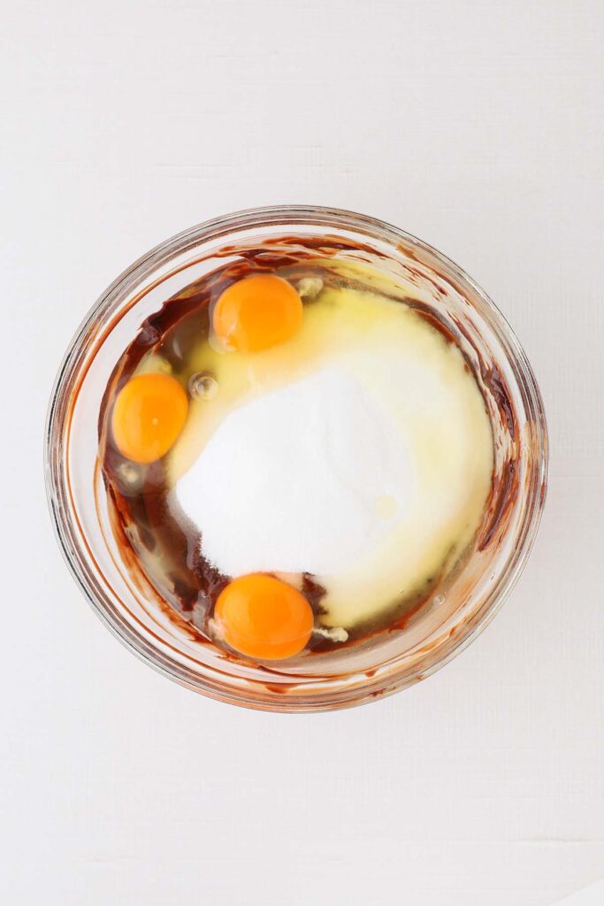 Eggs and granulated sugar added to melted chocolate in one bowl, showing the base for brownie cookie batter before mixing.