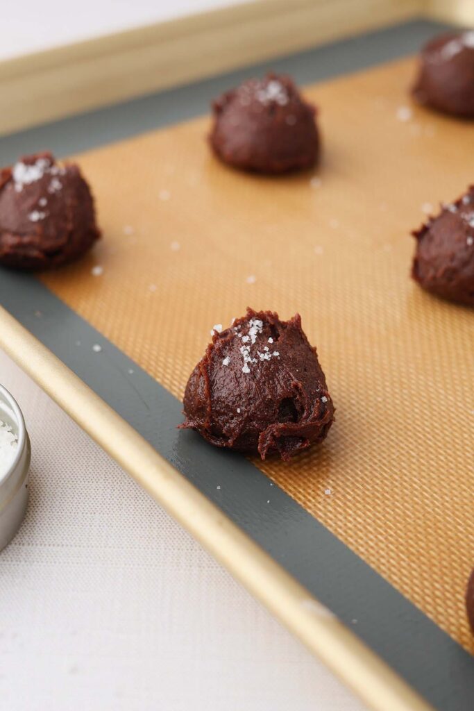 Scoops of brownie cookie dough topped with flaky salt on a lined baking sheet, showing texture before baking.