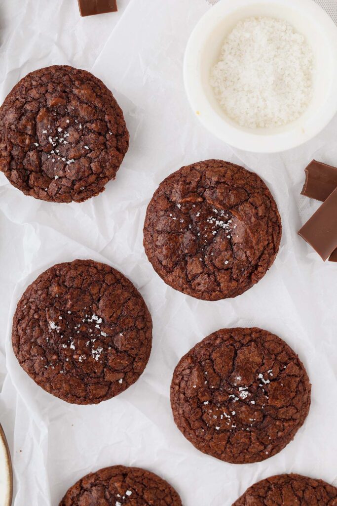 Fudgy brownie cookies with shiny crackly tops and a sprinkle of flaky salt, photographed overhead on parchment paper with chocolate pieces nearby.