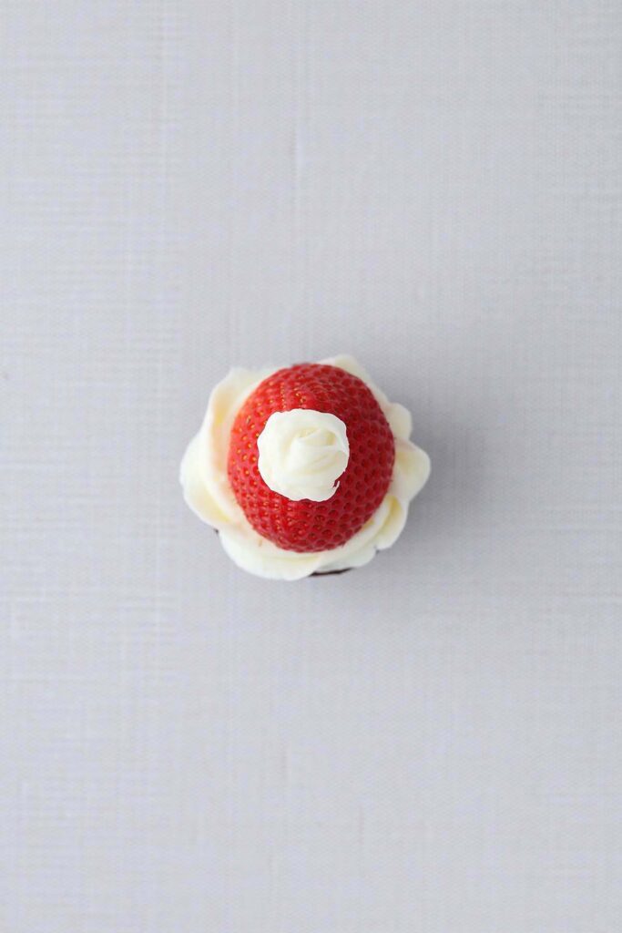 top down image showing a strawberry top side down sitting on white frosting with a white dot on top. This is on a white table top