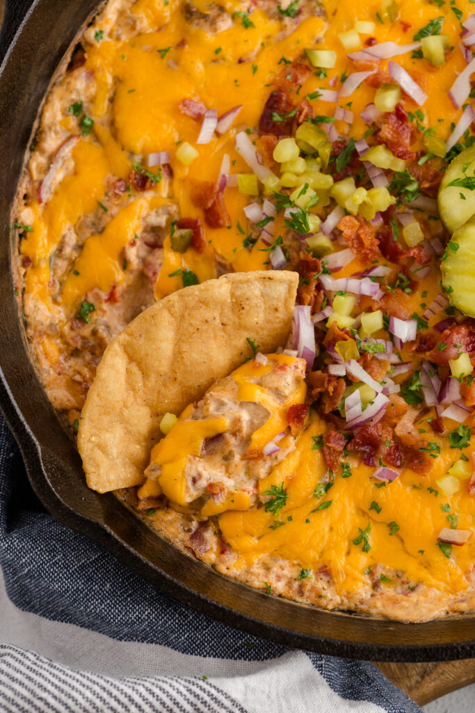 Cheeseburger dip baked in a cast iron skillet, topped with melted cheddar cheese, bacon, diced pickles, and red onion, served with tortilla chips on the side.