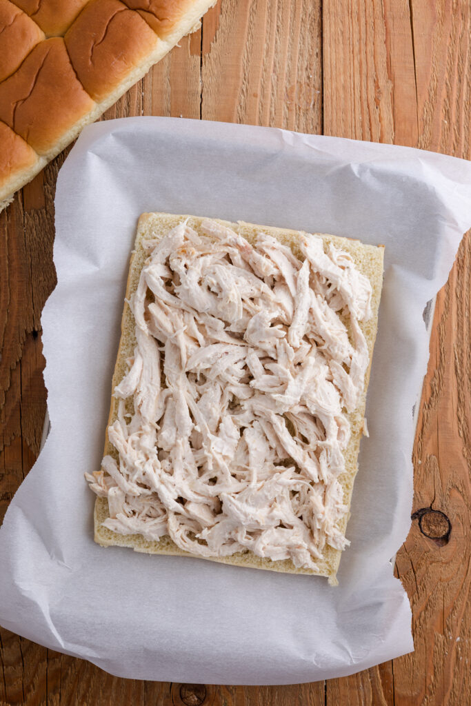 Shredded ranch chicken spread evenly over the bottom half of Hawaiian rolls in a lined baking dish