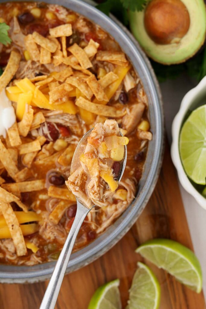 Close-up of chicken enchilada chili with shredded chicken, black beans, corn, and melted cheese, topped with tortilla strips, shown on a spoon with lime wedges nearby.