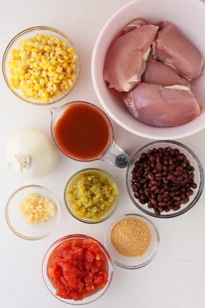 Ingredients for chicken enchilada chili laid out on a white surface, including raw chicken thighs, black beans, corn, enchilada sauce, diced tomatoes with green chilies, onion, garlic, taco seasoning, and diced green chilies.