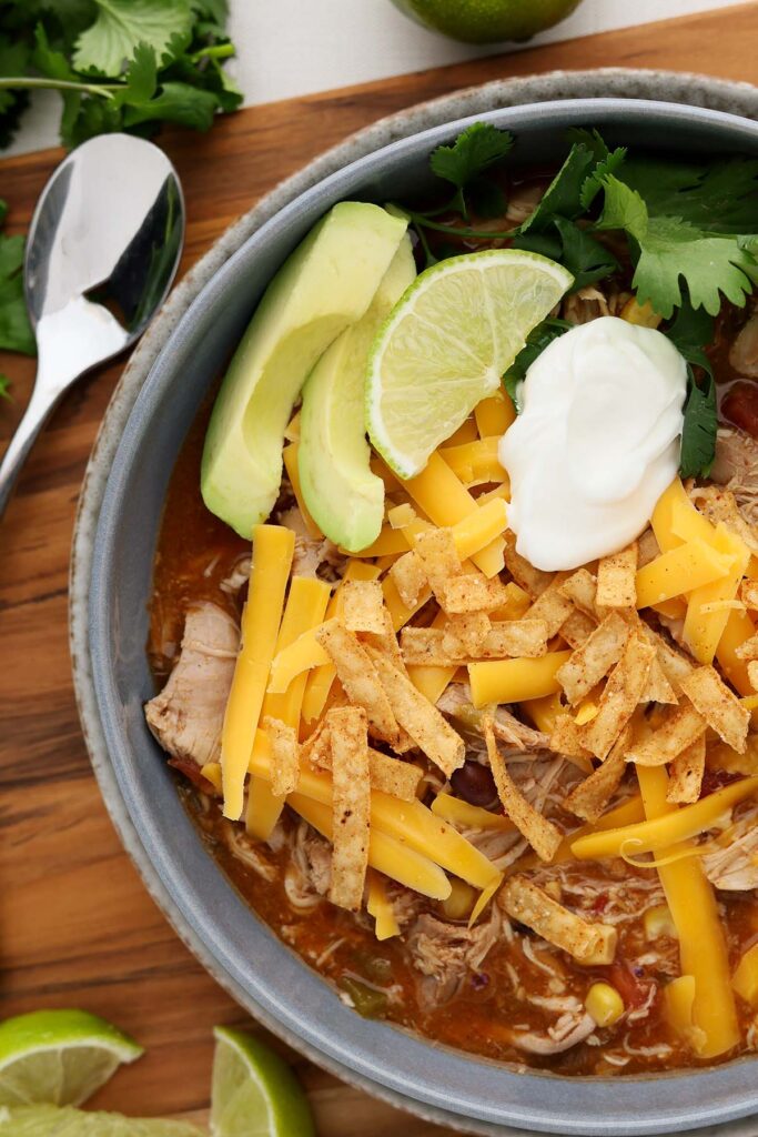 Top down image showing a super close up image of a bowl filled with a chicken chili that is topped with shredded cheese, sour cream, tortilla strips, lime, avocado, and sour cream
