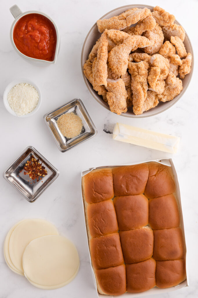 top down image showing chicken tenders, marinara, spices, butter, cheese, and rolls on a white table top