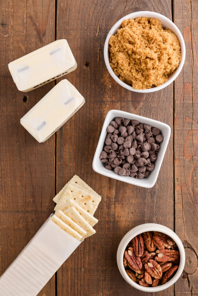 Ingredients for saltine cracker toffee laid out on a wooden surface, including butter sticks, a bowl of brown sugar, chocolate chips, saltine crackers, and chopped pecans.