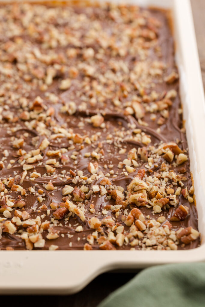 Baking sheet filled with freshly spread chocolate toffee topped with chopped pecans, cooling before being sliced into pieces.