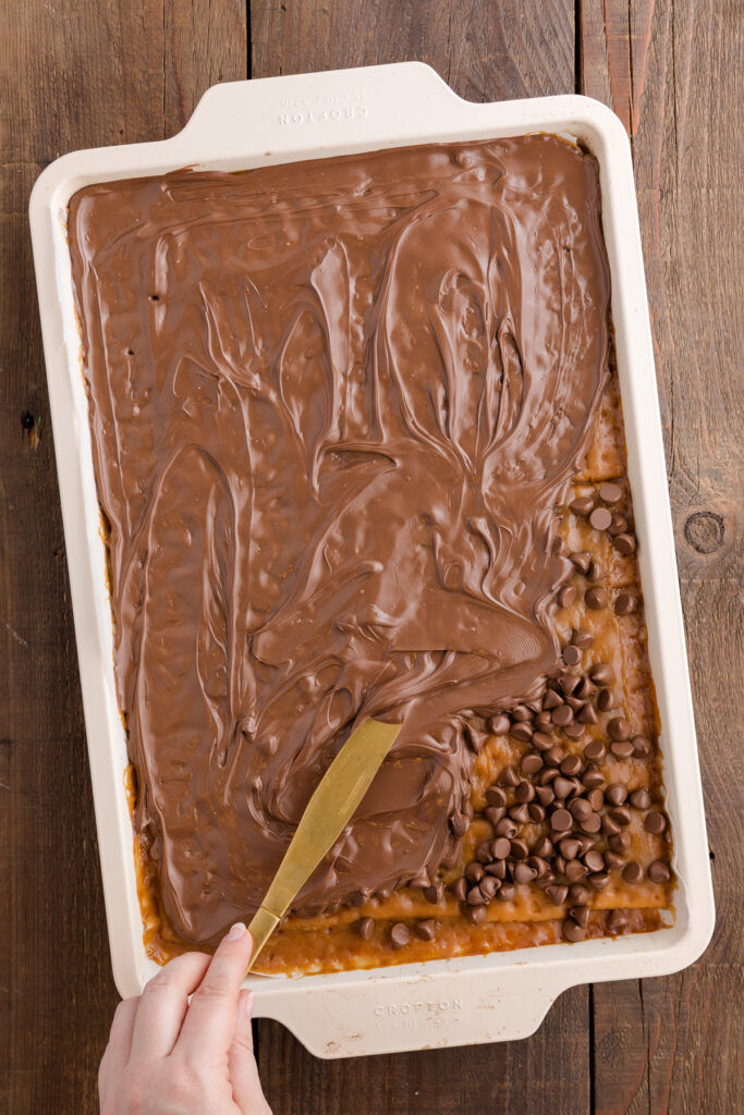 Baking sheet lined with saltine crackers arranged in a single even layer topped with toffee and sprinkled with chocolate chips that are being spread with a knife