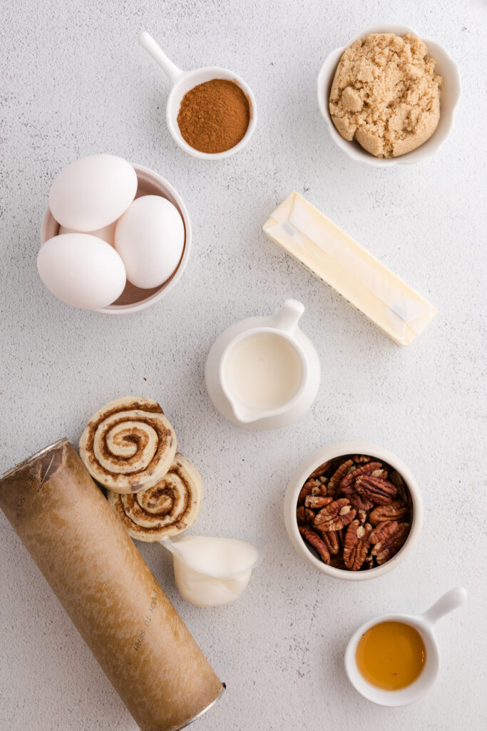top down image showing a can of cinnamon rolls, two cinnamon rolls and the frosting packet, pecans, syrup, cream, butter, eggs, cinnamon and brown sugar