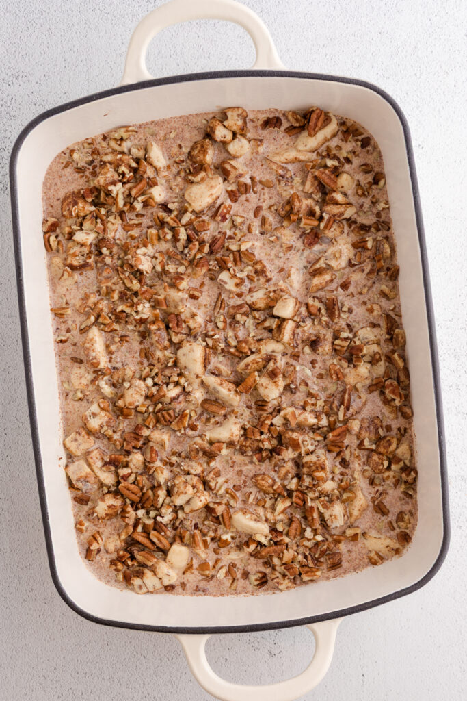 Top down image showing a 9x13 pan with a brown sugar mixture in the bottom topped with cut cinnamon rolls, cream, and pecans