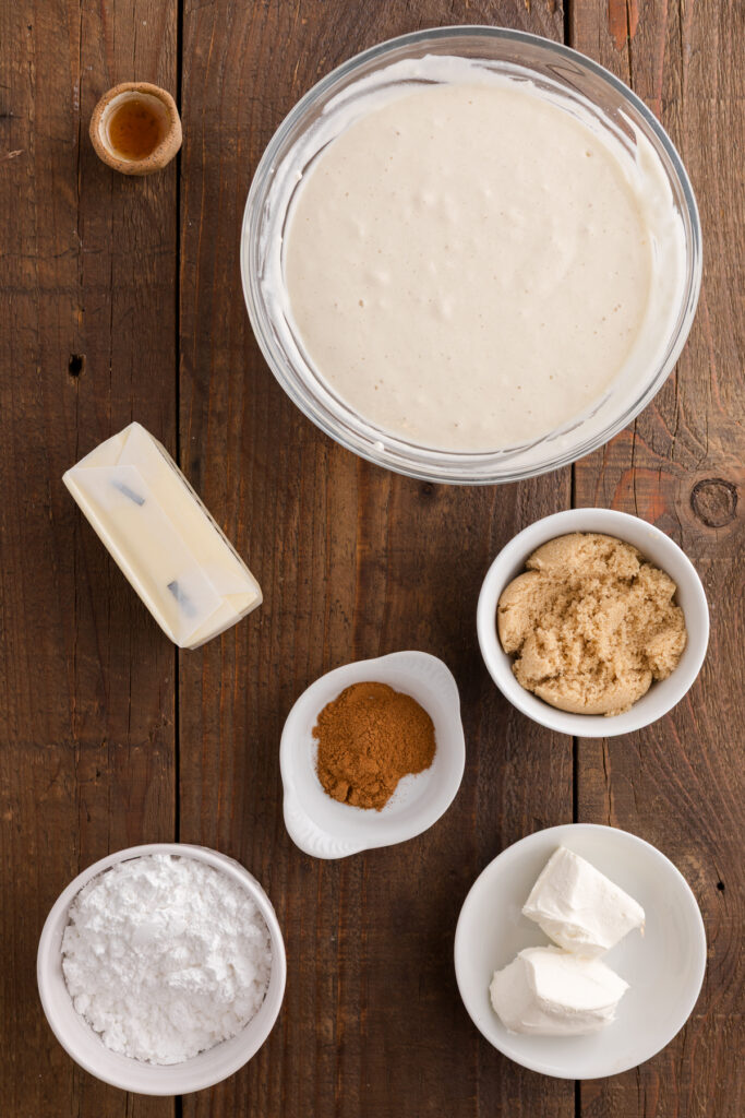 Overhead view of cream cheese frosting ingredients on a wooden surface, including cream cheese, butter, powdered sugar, brown sugar, cinnamon, vanilla, and a bowl of frosting.