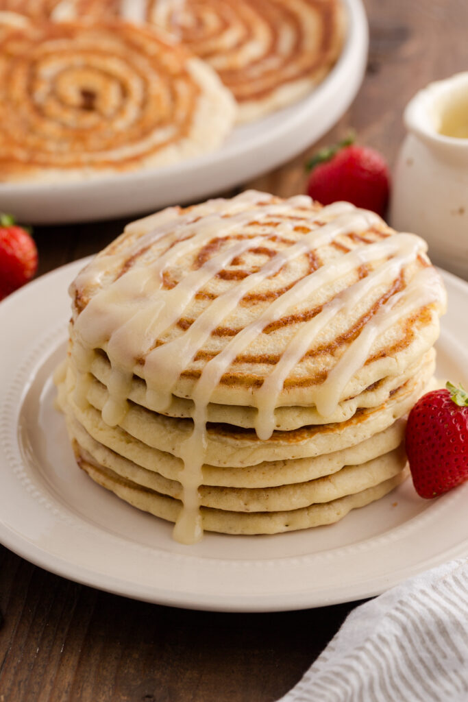 Stack of fluffy cinnamon roll pancakes drizzled with cream cheese glaze on a white plate, served with fresh strawberries and a pitcher of icing in the background.