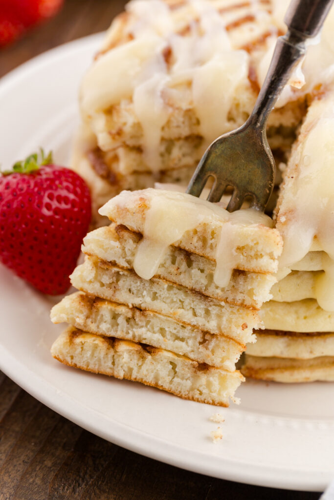 close up image showing a stack of pancakes with a cream cheese frosting with a bite on a fork. Strawberry in the background