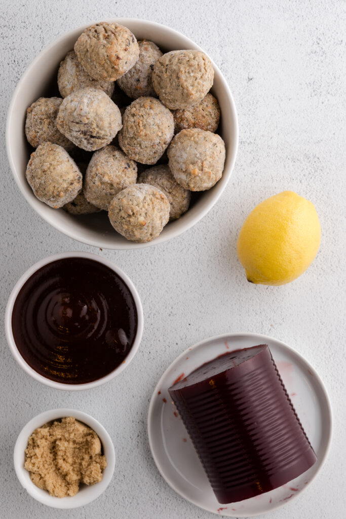 top down image showing a white table top with a bowl of meatballs, lemon, bbq sauce, canned cranberry sauce, and brown sugar