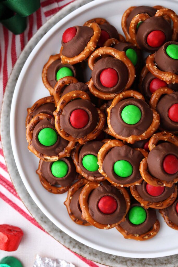 close up image showing a white round plate stacked with pretzels that are topped with chocolate with red and green M&m's pressed on top