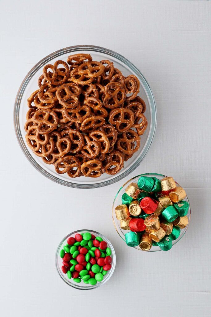 Top down image showing a white table with three glass bowls. One with pretzels, one with rolo candy,, one with MM