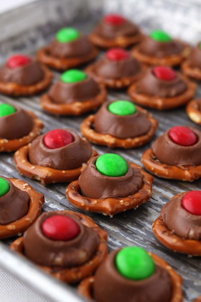 close up image showing a baking tray stacked with pretzels that are topped with chocolate with red and green M&m's pressed on top