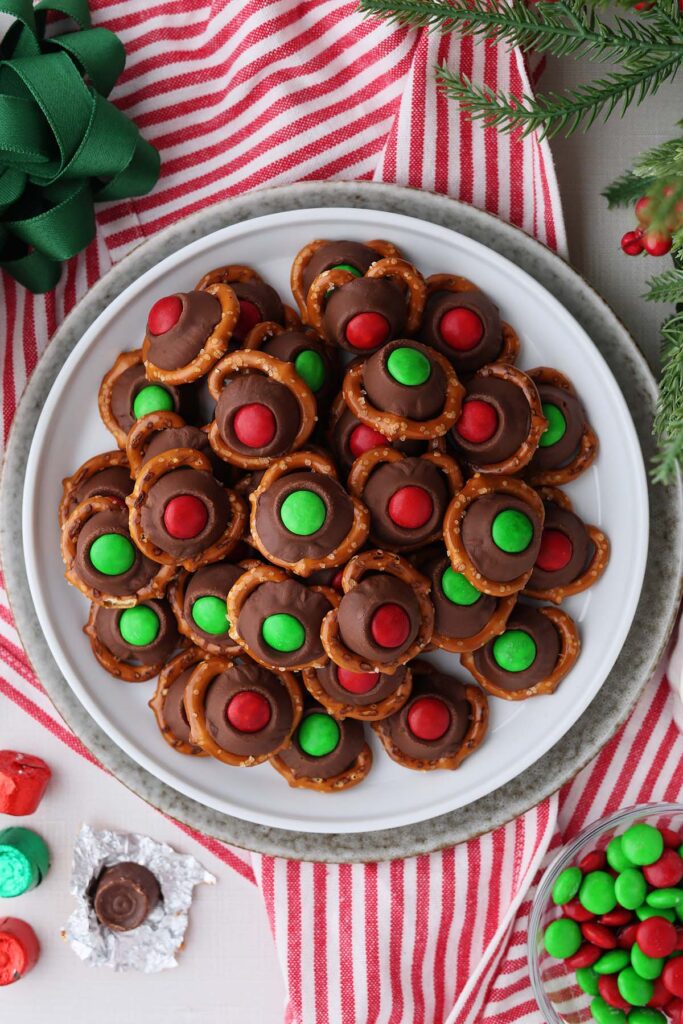 close up image showing a white round plate stacked with pretzels that are topped with chocolate with red and green M&m's pressed on top