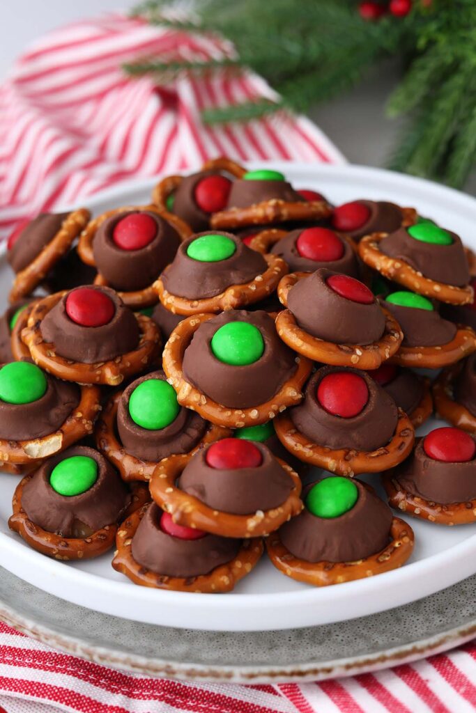 close up image showing a white round plate stacked with pretzels that are topped with chocolate with red and green M&m's pressed on top