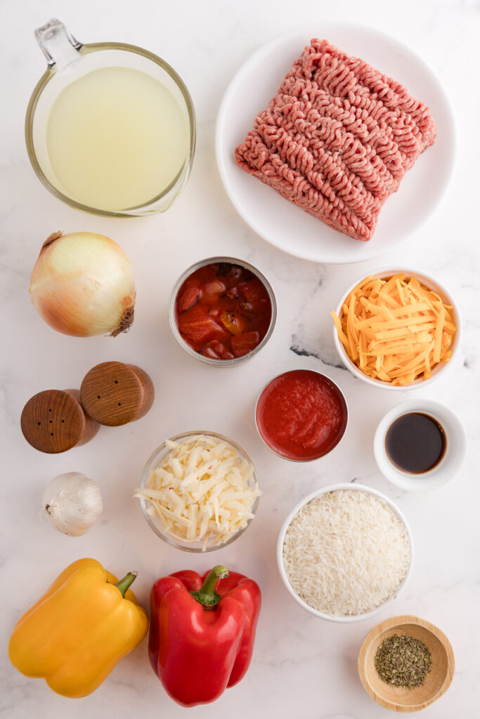 Overhead view of ingredients for a cheesy beef and rice casserole, including ground beef, uncooked rice, shredded cheese, bell peppers, onion, garlic, broth, diced tomatoes, tomato sauce, and seasonings on a white surface.