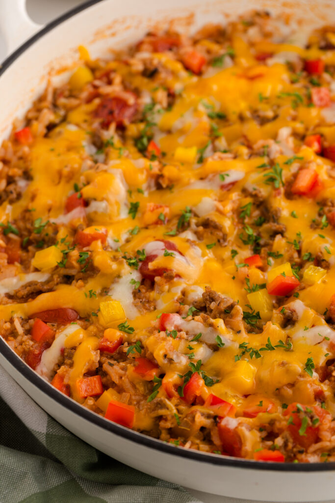 Close-up of a cheesy beef and rice casserole in a white skillet, with melted cheddar and mozzarella, diced red and yellow bell peppers, tomatoes, and fresh parsley on top.