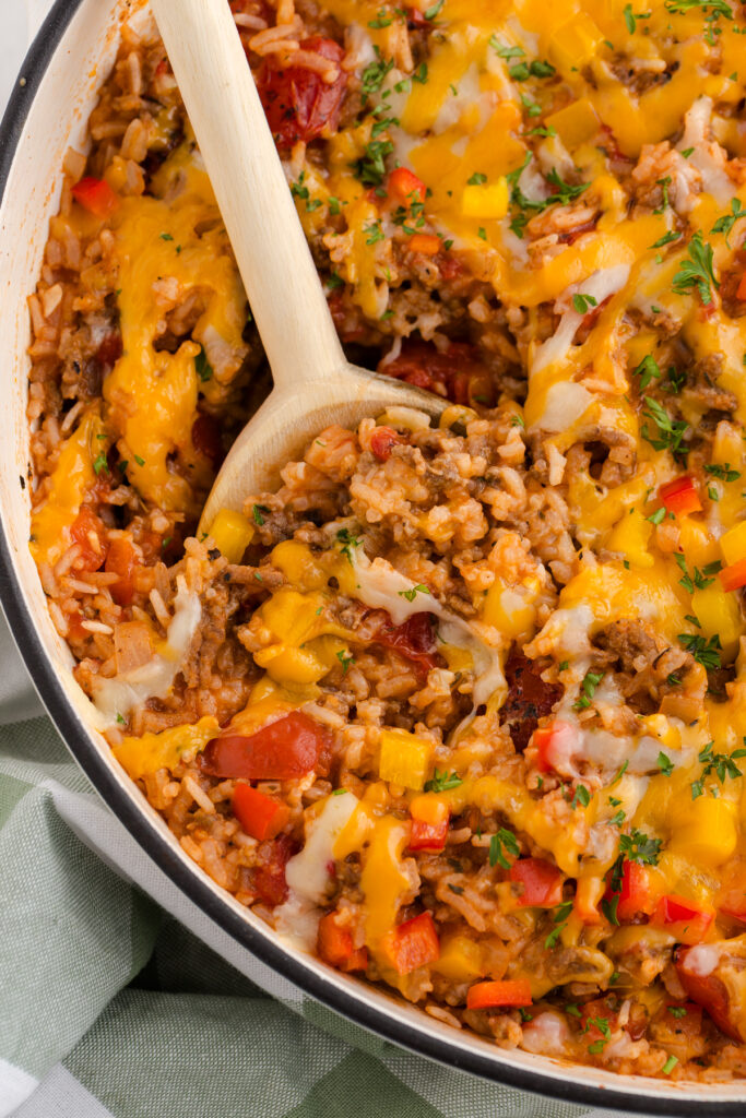 Close-up of a cheesy beef and rice casserole being scooped from a skillet, showing ground beef, tender rice, diced bell peppers, tomatoes, and melted cheese with fresh herbs.
