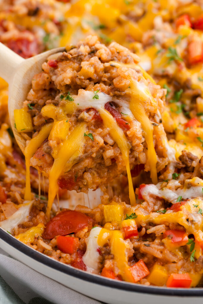 Cheesy beef and rice casserole being scooped from a baking dish with melted cheddar and mozzarella stretching, mixed with ground beef, rice, diced tomatoes, and bell peppers.