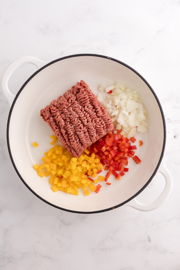 Raw ground beef added to a pot with chopped onion and diced red and yellow bell peppers before cooking.