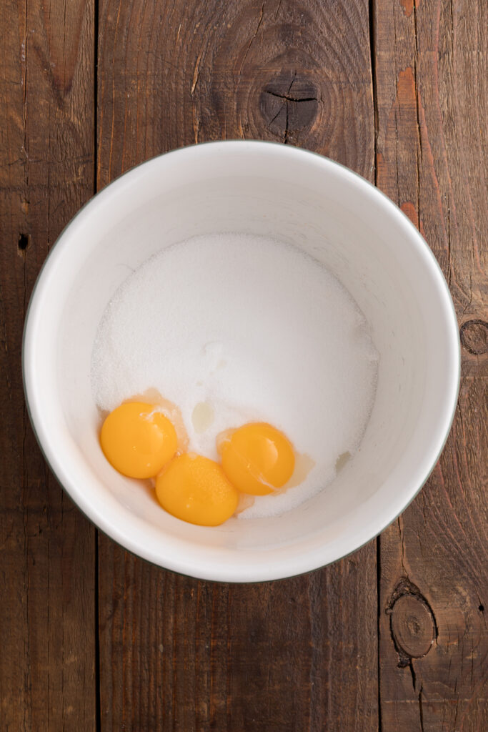 Top down image showing a white mixing bowl with sugar and egg yolks