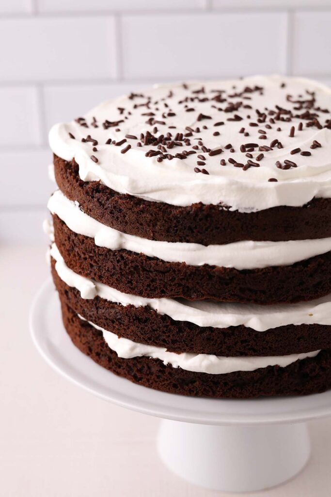 close up image showing 4 layers of chocolate cake with whipped cream in between sitting on a white cake plate with chocolate sprinkles on top