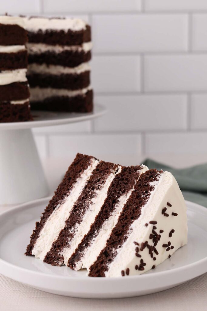 Chocolate Whipped Cream Cake Recipe a slice of chocolate layer cake with whipped cream and chocolate sprinkles sitting on a white plate with the full cake off in the background