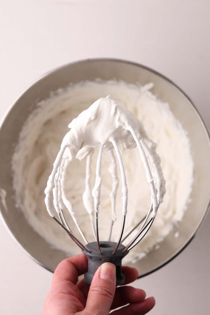 close up image of a hand holding the whisk attachment to a kitchenaid with whipped cream on it above the bowl