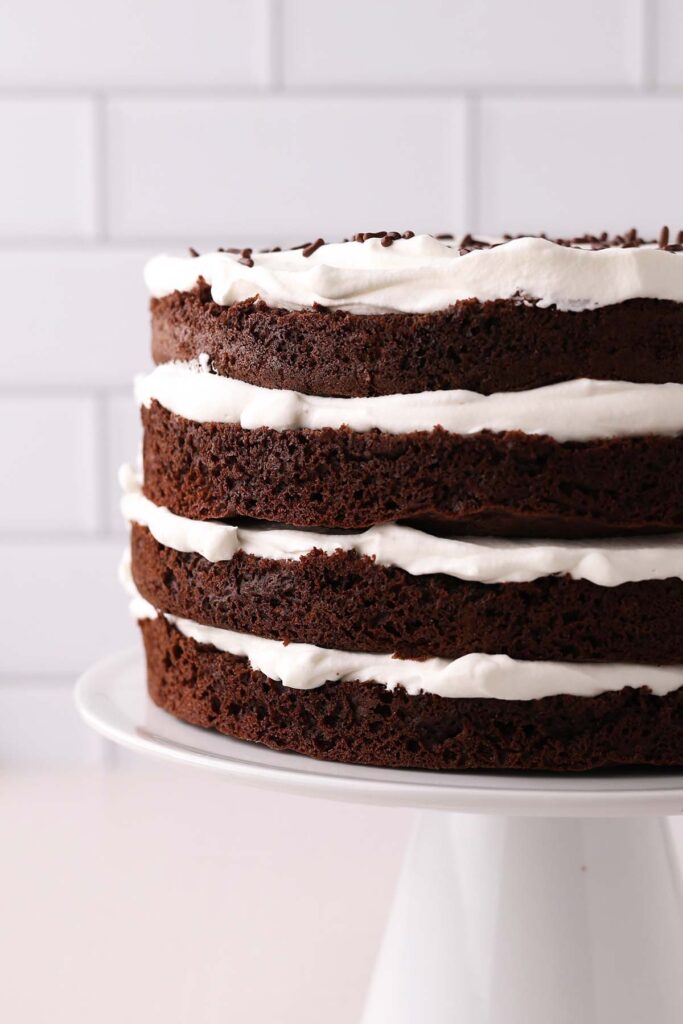 Whipped Cream Chocolate Cake close up image showing 4 layers of chocolate cake with whipped cream in between sitting on a white cake plate